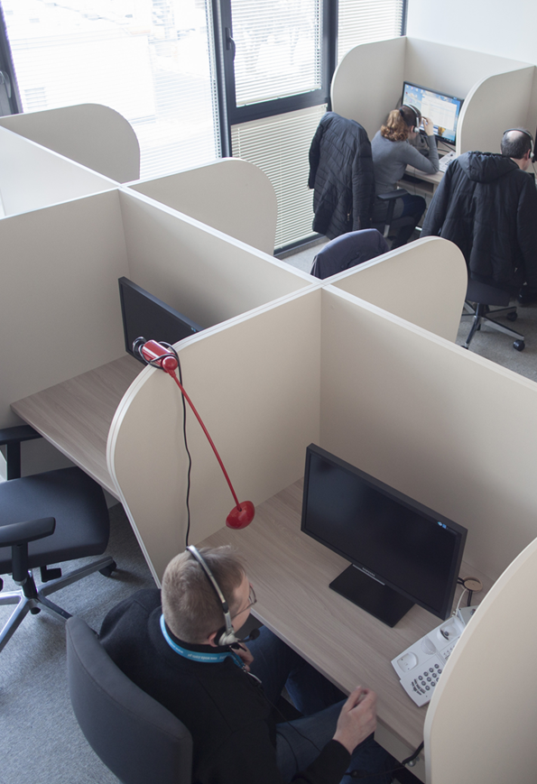 Stanowiska do pracy oddzielone ściankami działowymi. Przy biurkach siedzą pracownicy, ich twarze są niewidoczne, ujęcie pokazuje ich od tyłu. Na blatach ustawione są komputery i telefony.