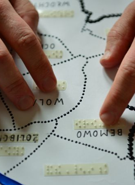 Dłonie studiujące mapę Warszawy z lokalizacjami zapisanymi alfabetem Braille'a.
